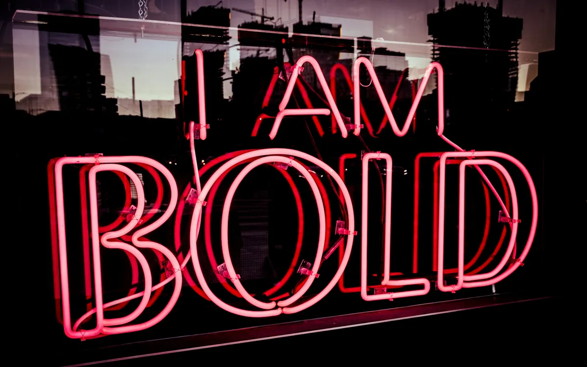 A pink neon sign reading 'I Am Bold' against a dark cityscape backdrop