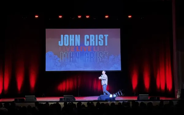 Comedian John Crist performing live on stage with a 'John Crist Live' screen behind him and the audience silhouetted in the foreground